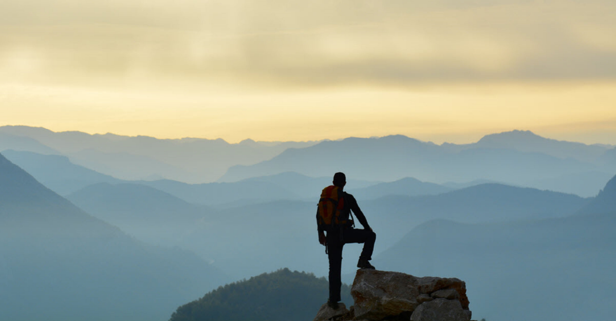 A hiker standing at the top of a mountain with one leg up on a rock, gazing out onto a landscape of hazy mountains and a golden sky