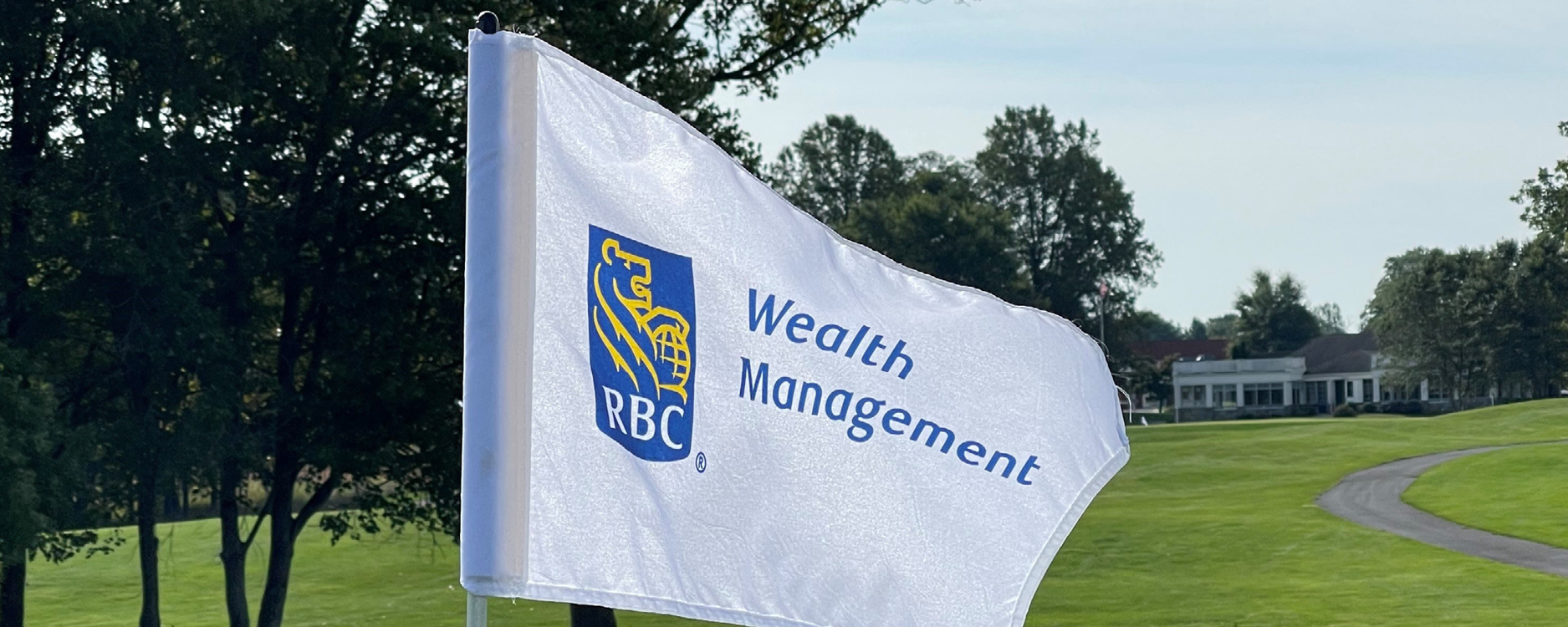 RBC Wealth Management pin flag waving in the breeze at Waverly Woods Golf Club with rolling grassy hills and clubhouse in distance
