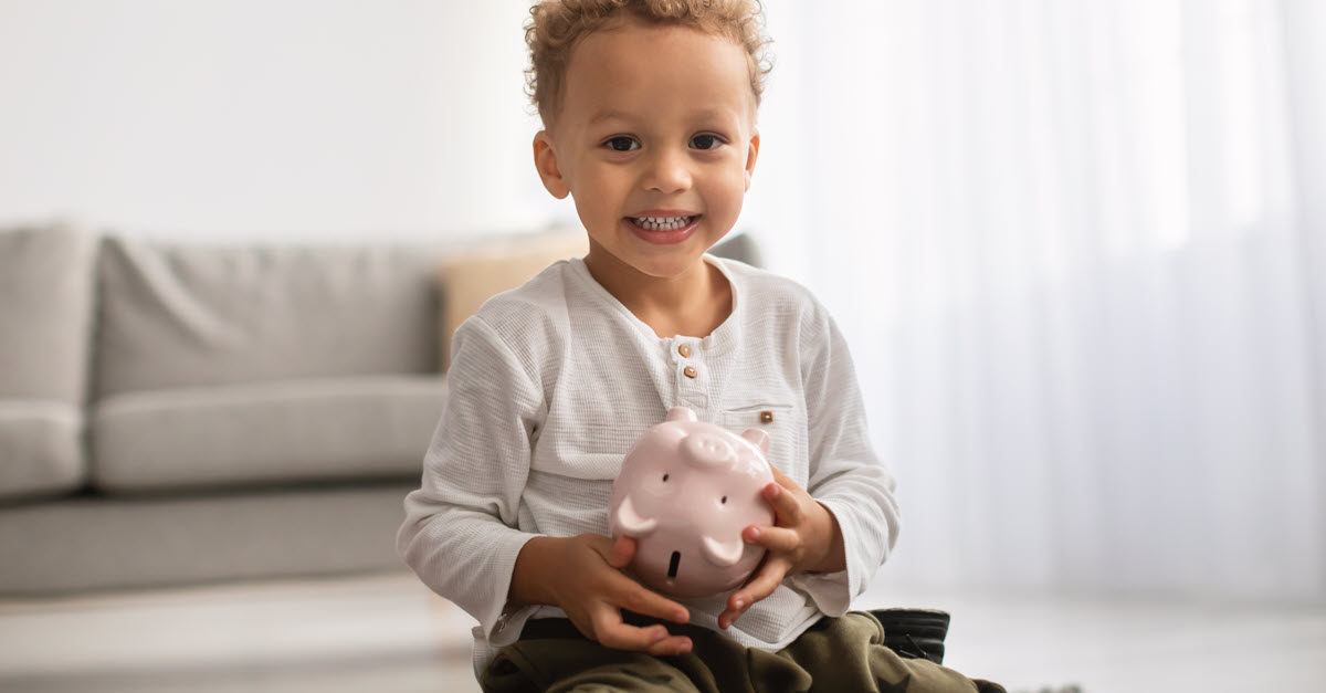 Smiling toddler holding a pink piggy bank while sitting on a rug in a bright living room
