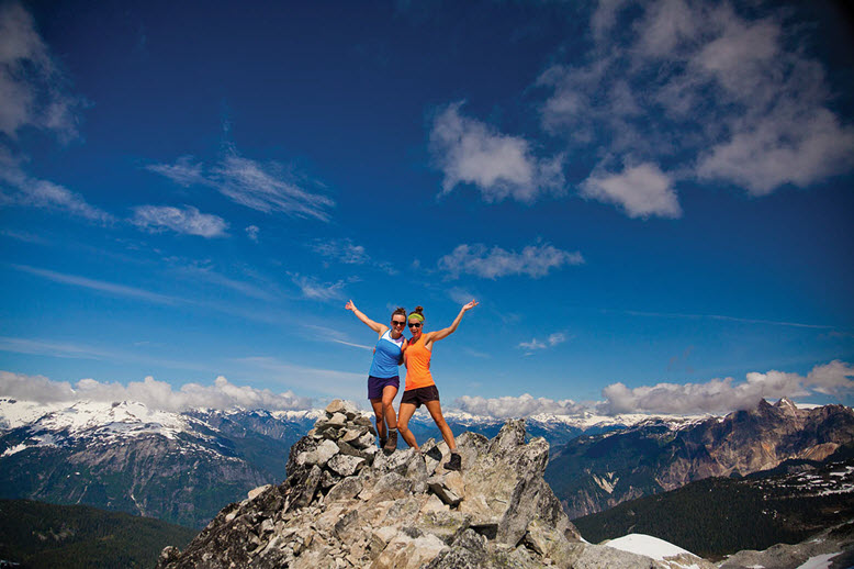 Two hikers standing at the top of a mountain with their arms raised in victory against a blue sky